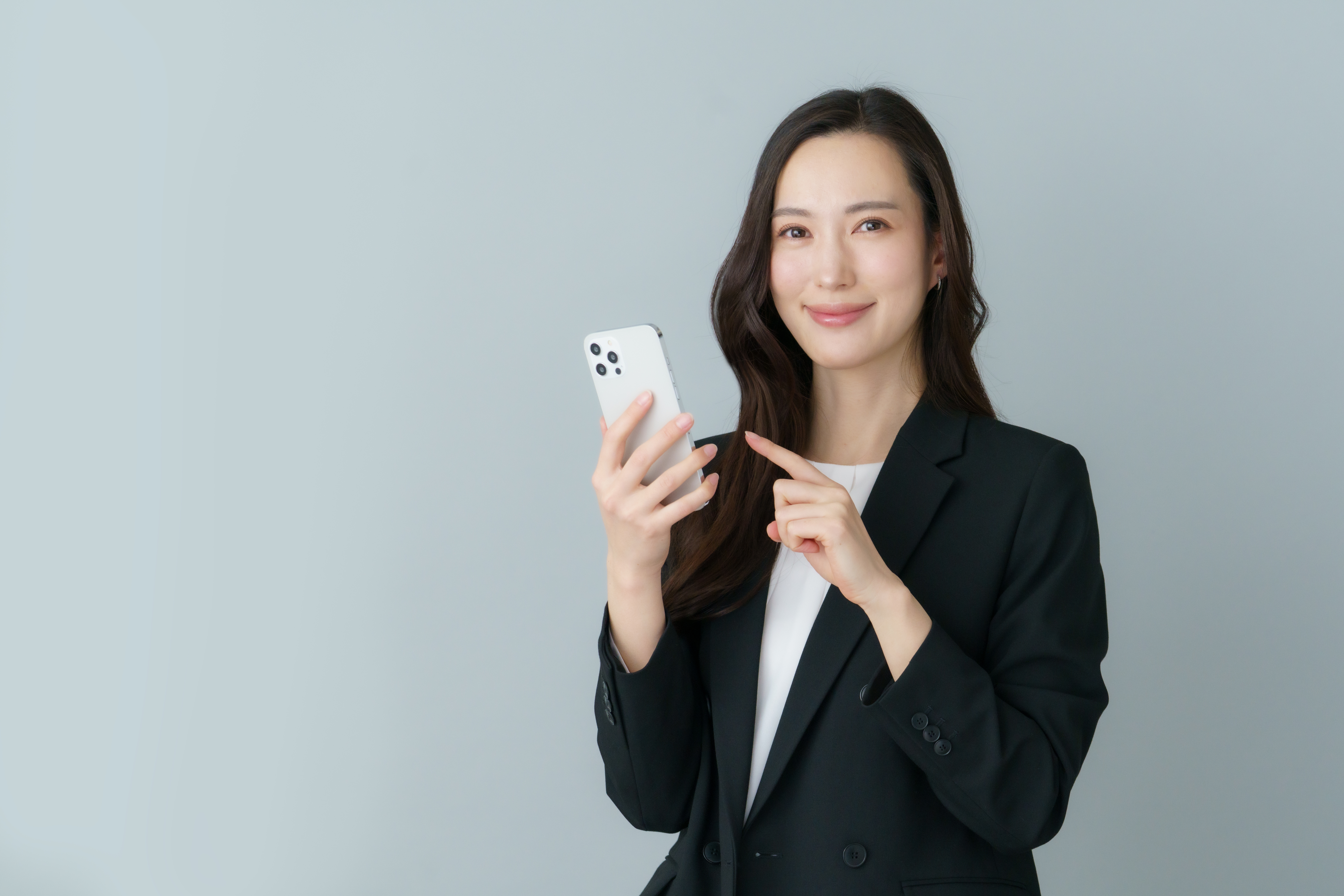 A young Japanese woman in a suit holding a smartphone with a gray background.